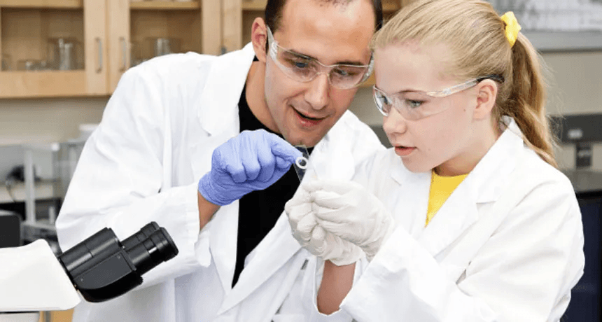 Mentor and student in lab coats and goggles reviewing a microscope slide together at a lab bench