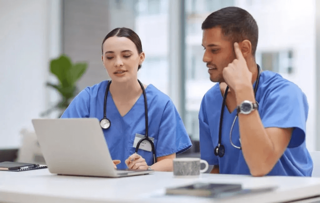 Two healthcare professionals in scrubs collaborating at a laptop in a clinical learning setting
